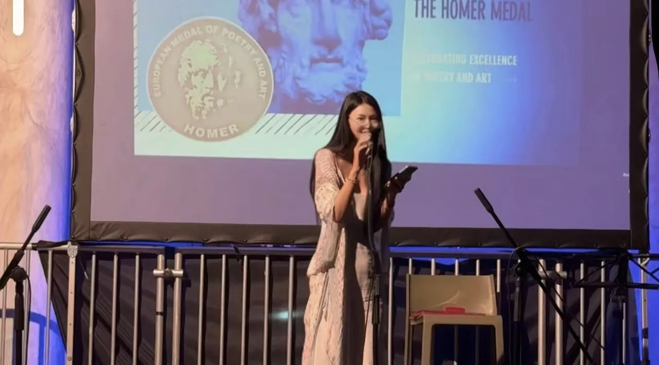 Sula reading poetry on stage at the Genoa International Poetry Festival, Homer Medal ceremony in the background